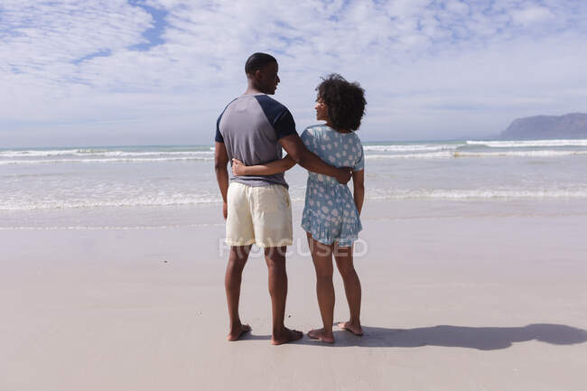 Африканская американская пара стоит и обнимается на пляже улыбаясь. здоровый отдых на открытом воздухе у моря. — стоковое фото