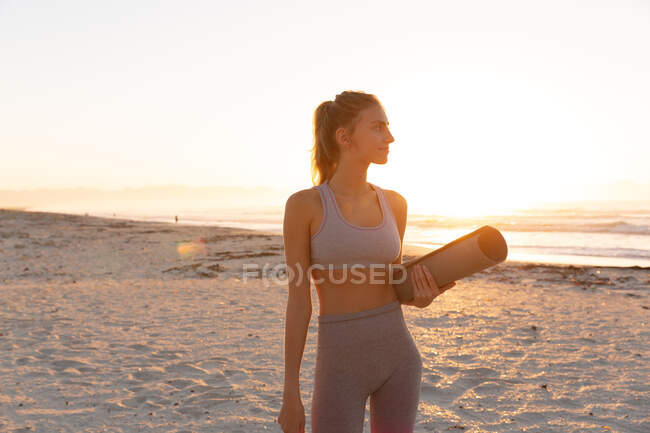 Mujer caucásica sosteniendo esterilla de yoga sonriendo mientras está de pie en la playa. Yoga fitness y concepto de estilo de vida saludable - foto de stock