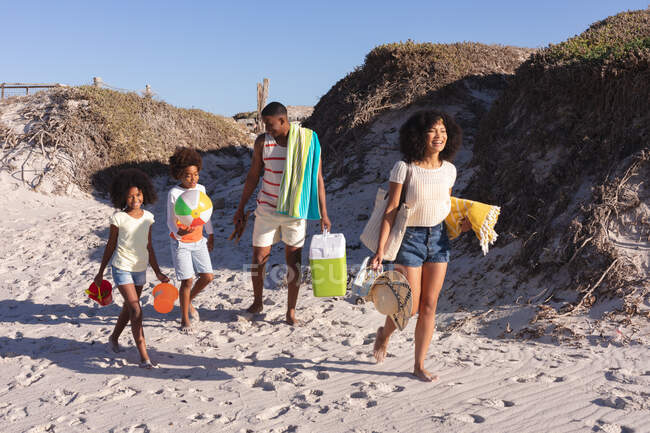 Африканские американские родители и двое детей с пляжными аксессуарами прогуливаются по пляжу. семейное свободное время у моря. — стоковое фото