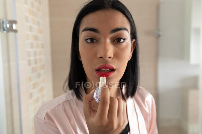 Retrato de mujer transgénero de raza mixta reflejado en el espejo del baño poniéndose lápiz labial. permanecer en casa aislado durante el bloqueo de cuarentena. - foto de stock