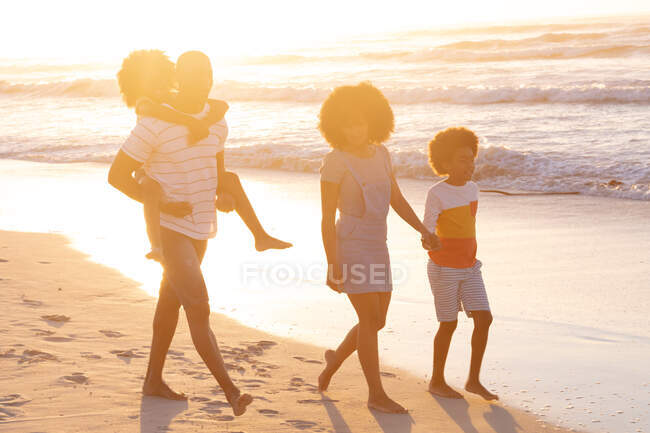 Батьки-афроамериканці і двоє дітей ходять, тримаючи за руки і тримаючись за свиней на пляжі. Здоровий вільний час на відкритому повітрі біля моря. — стокове фото
