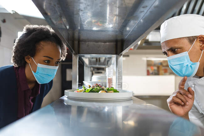 Mixed race professional chef and kitchen manager wearing face masks looking at finished dish. working in a busy restaurant kitchen during coronavirus covid 19 pandemic. — Stock Photo