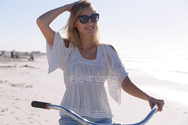 Улыбающаяся белая женщина в солнечных очках, в белом топе и в шортах, сидящая на велосипеде на пляже. здоровый отдых на открытом воздухе у моря. — стоковое фото