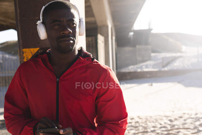 Uomo afroamericano che si allena all'aperto, indossa le cuffie, usa lo smartphone sotto il ponte. sano stile di vita all'aperto allenamento fitness. — Foto stock