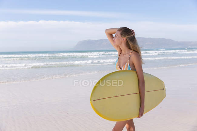 Mujer caucásica con bikini llevando tabla de surf en la playa. tiempo de ocio al aire libre saludable junto al mar. - foto de stock