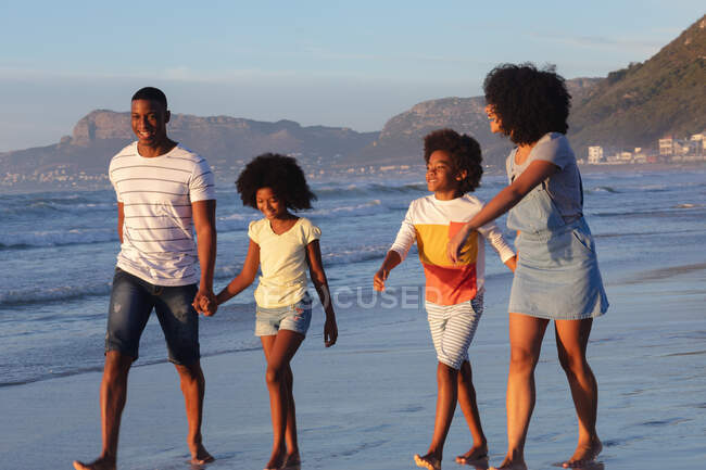 Улыбающиеся африканские американские родители и двое детей, идущих и держащихся за руки на пляже. здоровый отдых на открытом воздухе у моря. — стоковое фото