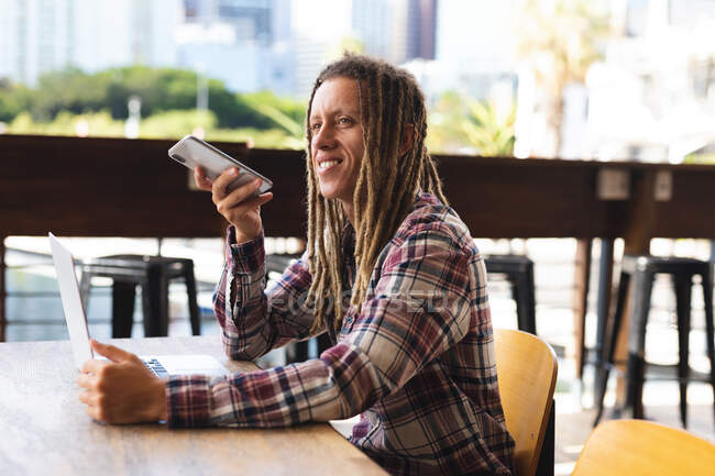 Lächelnder, gemischter Mann mit Dreadlocks, der am Tisch vor dem Café sitzt und mit dem Smartphone spricht. digitaler Nomade, unterwegs in der Stadt. — Stockfoto