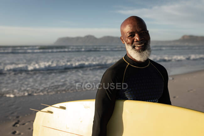 Retrato de un hombre afroamericano mayor sosteniendo una tabla de surf sonriendo en la playa. viaje vacaciones retiro estilo de vida concepto - foto de stock
