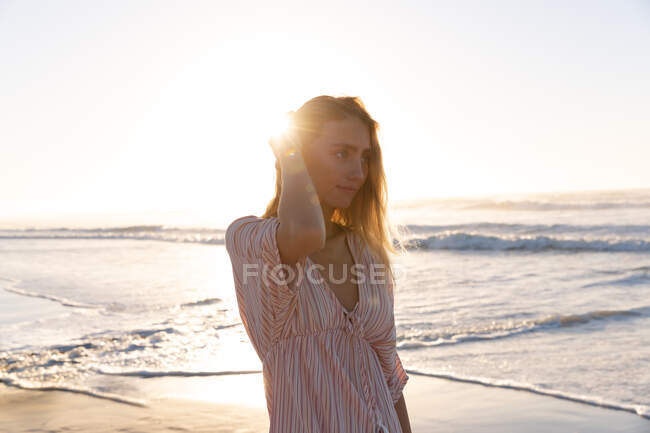 Красива біла жінка торкається волосся, що стоїть на пляжі під час заходу сонця. концепція літнього пляжного відпочинку . — стокове фото