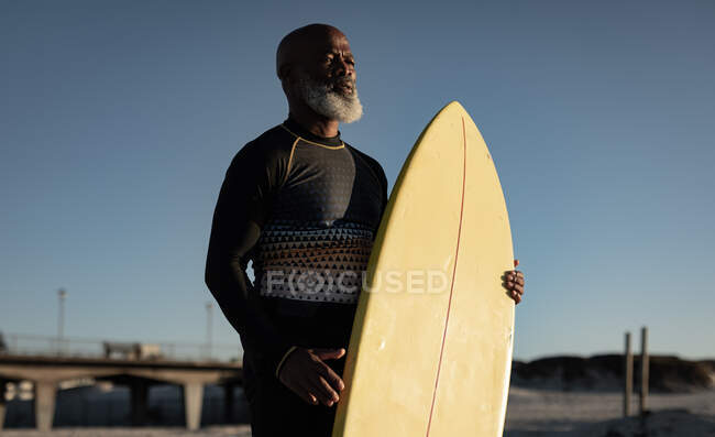 Ein älterer afrikanisch-amerikanischer Mann mit Surfbrett steht am Strand. Reise Urlaub Ruhestand Lifestylekonzept — Stockfoto