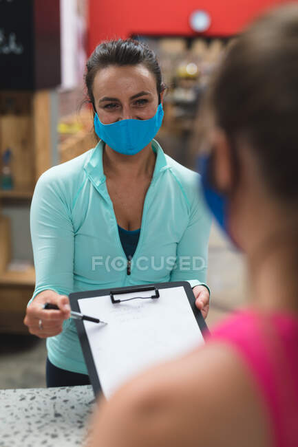 Femme caucasienne portant un masque facial derrière le comptoir à la réception de la salle de gym passant document à chanter. fitness et temps libre à la salle de gym pendant coronavirus covid 19 pandémie. — Photo de stock