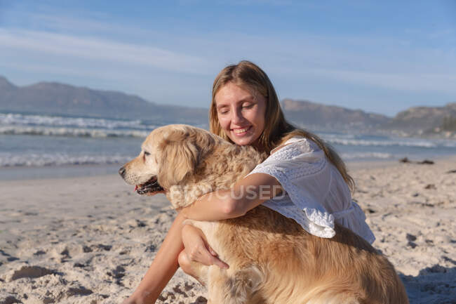 Mujer caucásica sentada sobre arena abrazando a un perro en la playa. tiempo de ocio al aire libre saludable junto al mar. - foto de stock