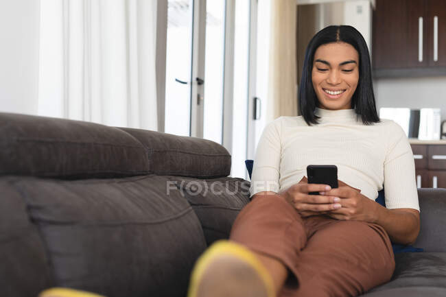 Glückliche Transgender-Frau mit gemischter Rasse, die es sich im Wohnzimmer gemütlich macht und auf der Couch Selfies macht. Isolationshaft während der Quarantäne. — Stockfoto