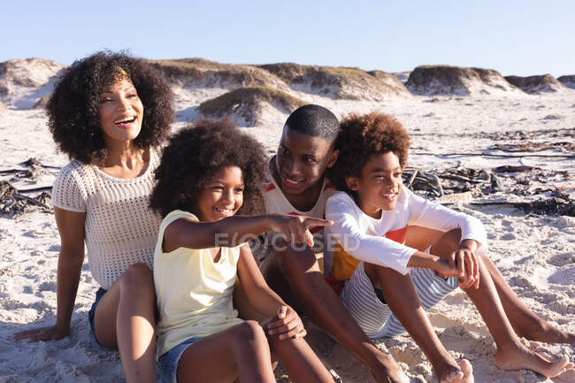 Африканські американські батьки і двоє дітей сидять і сміються на пляжі. сім'я на відкритому повітрі відпочиває біля моря. — стокове фото