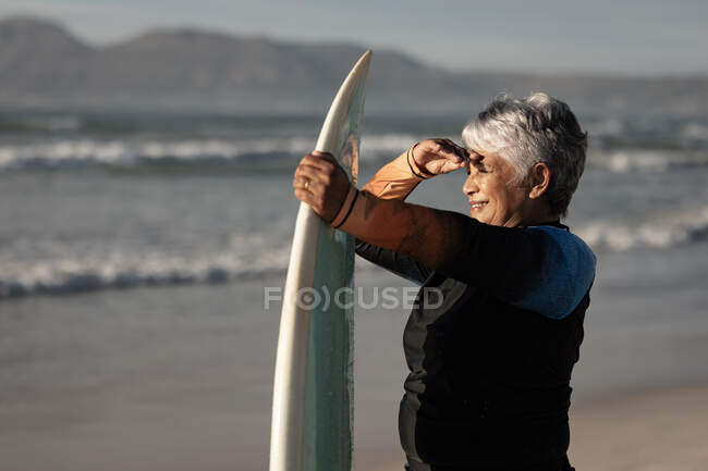 Mulher americana africana sênior com prancha de surf olhando para uma distância na praia. viagem férias aposentadoria estilo de vida conceito — Fotografia de Stock