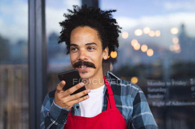Fröhlicher, gemischter männlicher Barista mit Schnurrbart, der in der Tür eines Cafés lehnt und mit dem Smartphone spricht. unabhängiges Kleinunternehmen in einer Stadt. — Stockfoto