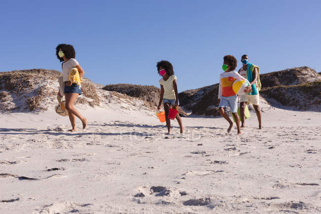 Genitori afroamericani e due bambini che tengono in mano accessori da spiaggia passeggiando in spiaggia. famiglia tempo libero all'aperto al mare durante coronavirus covid 19 pandemia. — Foto stock