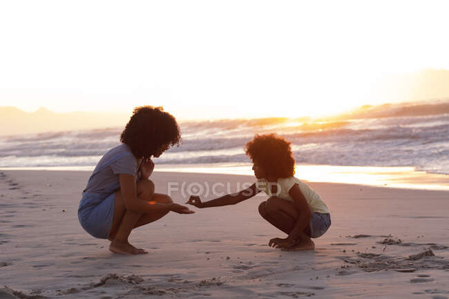 African american mother and daughter collecting shells at the beach smiling. healthy outdoor leisure time by the sea. - foto de stock