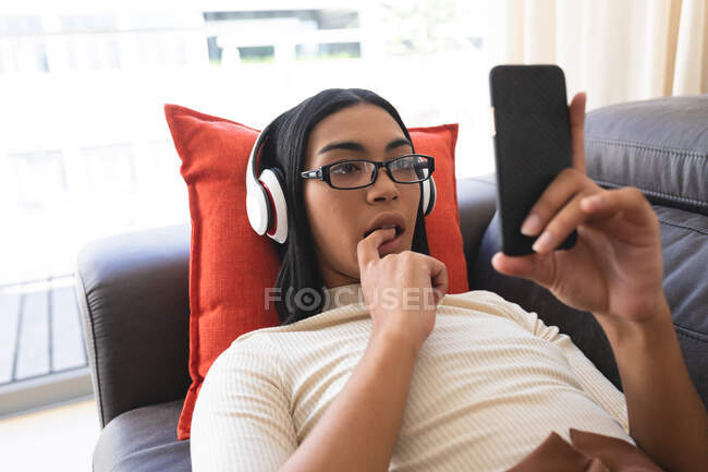 Mujer transexual de raza mixta relajándose tumbada en el sofá con auriculares usando un teléfono inteligente. permanecer en casa aislado durante el bloqueo de cuarentena. - foto de stock