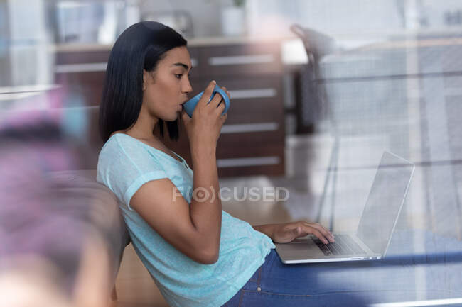 Mujer transgénero de raza mixta que trabaja en casa usando una computadora portátil bebiendo café. permanecer en casa aislado durante el bloqueo de cuarentena. - foto de stock