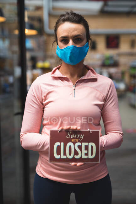 Porträt einer kaukasischen Frau mit Maske und offenem Schild auf dem Flur eines Fitnessstudios. Fitness und Freizeit im Fitnessstudio während der Coronavirus-Pandemie 19. — Stockfoto