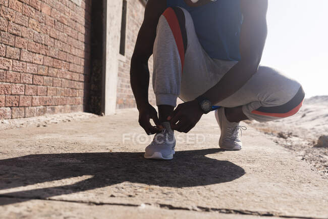 Unterteil eines afrikanisch-amerikanischen Mannes, der an sonnigen Tagen Schnürsenkel bindet. gesundes Outdoor-Fitness-Training. — Stockfoto