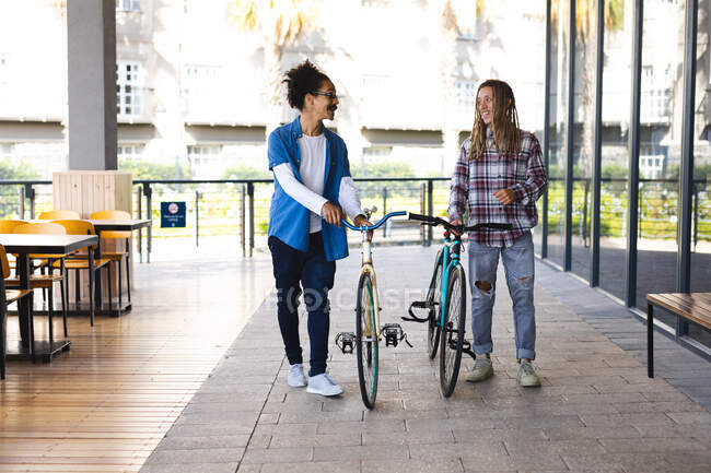 Дві щасливі змішані раси друзі чоловічої статі ковзають на велосипедах на вулиці і розмовляють. зелений міський спосіб життя, на вулиці і близько в місті . — стокове фото