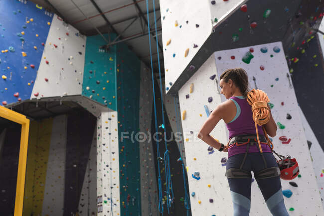 Femme caucasienne avec une corde au-dessus de son épaule se préparant à grimper au mur d'escalade intérieur. fitness et temps libre au gymnase. — Photo de stock