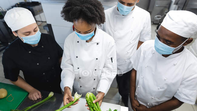 Diversi chef professionisti di razza maschile e femminile che preparano verdure indossando maschere per il viso. lavorando in una cucina ristorante occupato durante coronavirus covid 19 pandemia. — Foto stock