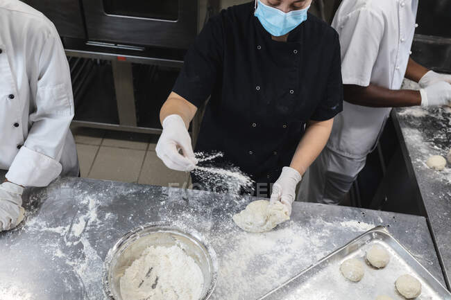 Gemischte Profiköche bereiten Teig mit Hygienehandschuhen und Mundschutz zu. Arbeit in einer belebten Restaurantküche während der Coronavirus-Pandemie 19. — Stockfoto