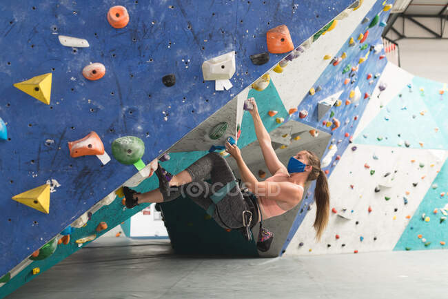 Mujer caucásica con máscara trepando por una pared en el gimnasio de escalada interior. fitness y tiempo libre en el gimnasio durante coronavirus covid 19 pandemia. - foto de stock