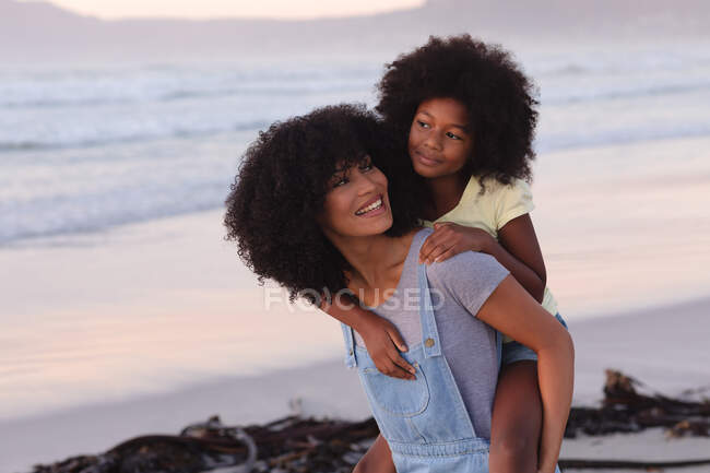 Smiling african american mother carrying her daughter piggyback at the beach. healthy outdoor leisure time by the sea. — Stock Photo