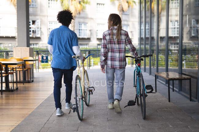 Two mixed race male friends wheeling bicycles in the street and talking. green urban lifestyle, out and about in the city. — Stock Photo