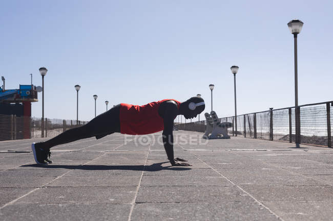 Homem afro-americano se exercitando, usando fones de ouvido no cais no dia ensolarado. treinamento de fitness ao ar livre saudável. — Fotografia de Stock