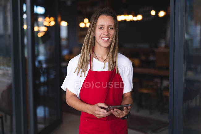 Portrait of happy mixed race male barista with dreadlocks standing in doorway of cafe holding tablet. independent small business in a city. — Stock Photo