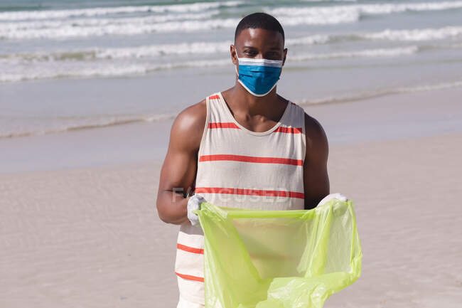 African american man wearing face mask collecting rubbish from the beach. eco beach conservation during coronavirus covid 19 pandemic. - foto de stock