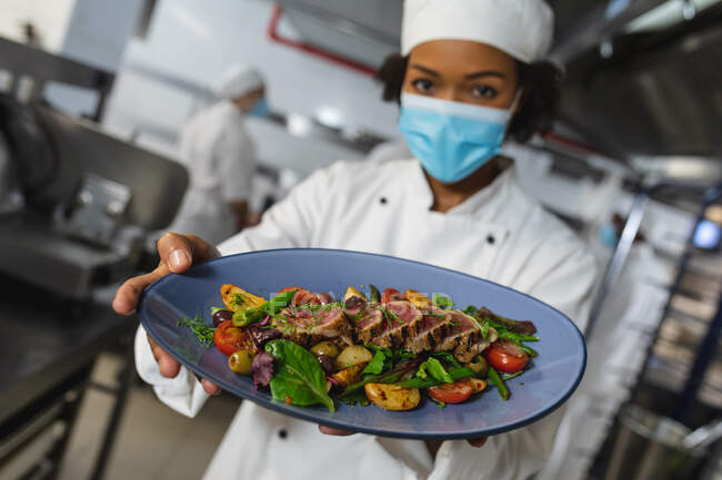 Mixed race professional chef with finished dish before serving wearing face mask. working in a busy restaurant kitchen during coronavirus covid 19 pandemic. — Stock Photo