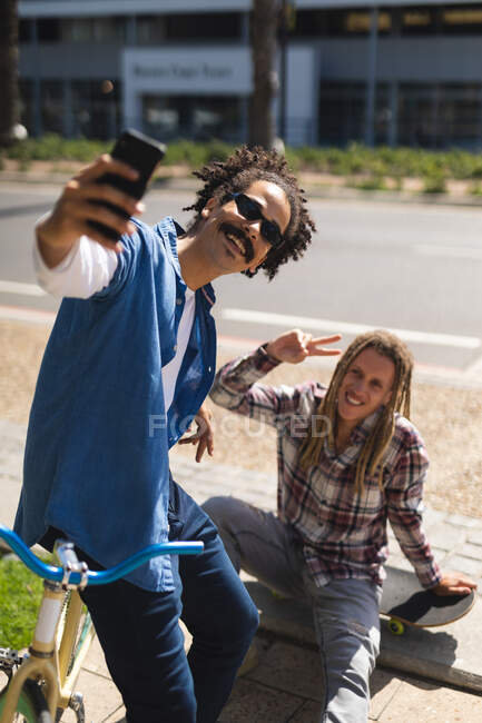 Dos felices amigos varones de raza mixta sentados en monopatín y en bicicleta en la calle y tomando selfie. nómada digital, fuera y alrededor de la ciudad. - foto de stock