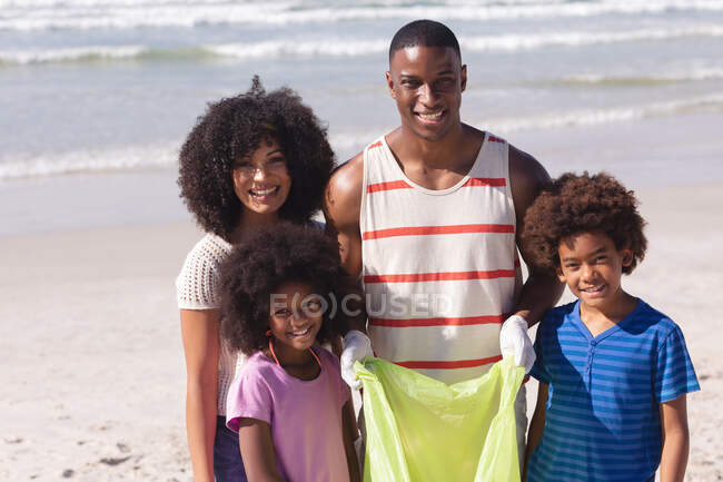 Afrikanische amerikanische Eltern mit zwei Kindern, die lächelnd Müll am Strand sammeln. Öko-Strandschutz für Familien. — Stockfoto