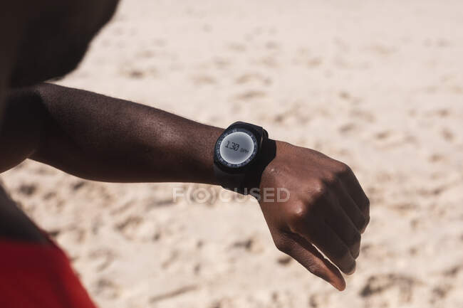 African american man exercising, looking at smartwatch on beach on sunny day. healthy outdoor lifestyle fitness training. — Stock Photo