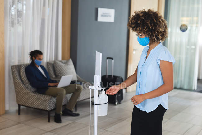 Mujer de negocios de raza mixta desinfectando manos en el vestíbulo del hotel. viaje de negocios hotel durante coronavirus covid 19 pandemia. - foto de stock
