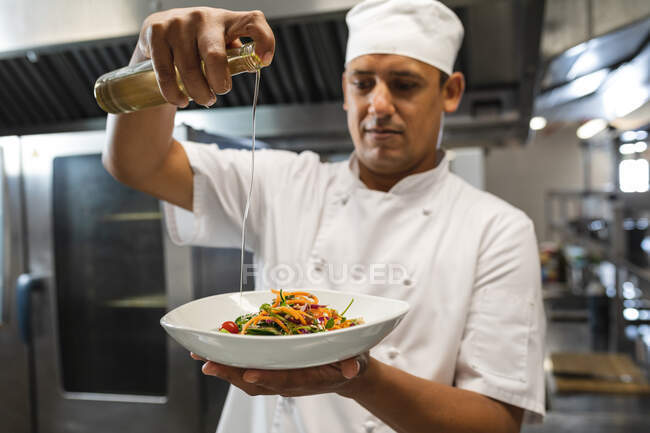 Mixed race male professional chef finishing dish before serving. working in a busy restaurant kitchen. — Stock Photo