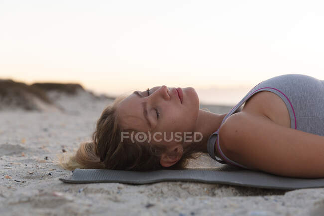Femme caucasienne méditant alors qu'elle était allongée sur un tapis de yoga à la plage. yoga fitness et mode de vie sain concept — Photo de stock