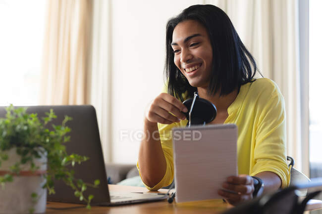 Mujer transgénero de raza mixta feliz haciendo videollamadas usando laptop, sonriendo, sosteniendo notas. permanecer en casa aislado durante el bloqueo de cuarentena. - foto de stock