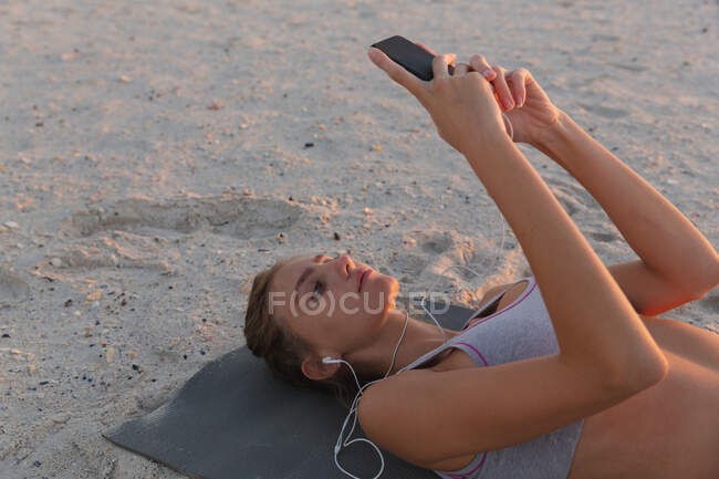 Femme caucasienne portant des écouteurs à l'aide d'un smartphone tout en étant allongée sur un tapis de yoga à la plage. yoga fitness et mode de vie sain concept — Photo de stock