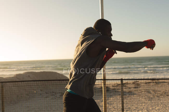 Afroamerikanischer Mann beim Schattenboxen im Freien mit eingewickelten Händen am Strand bei Sonnenuntergang. gesundes Outdoor-Fitness-Training. — Stockfoto