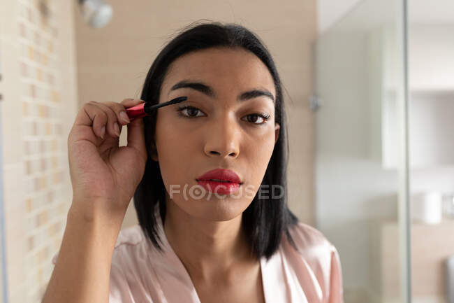 Retrato de mulher mista transgênero refletida no espelho do banheiro colocando rímel. ficar em casa em isolamento durante o confinamento de quarentena. — Fotografia de Stock