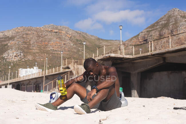 African american man exercising, resting on beach, holding bottle on sunny day. healthy outdoor lifestyle fitness training. — Foto stock