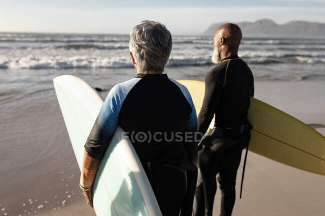 Casal sénior diverso na praia segurando pranchas de surf olhando para o mar. saúde e bem-estar, reforma activa. — Fotografia de Stock