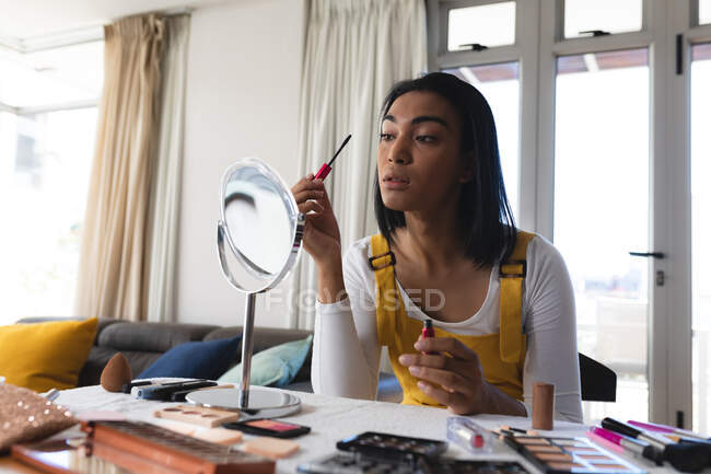 Transgender-Frau mit gemischter Rasse sitzt am Tisch und schaut in den Spiegel und setzt sich Mascara auf. Isolationshaft während der Quarantäne. — Stockfoto